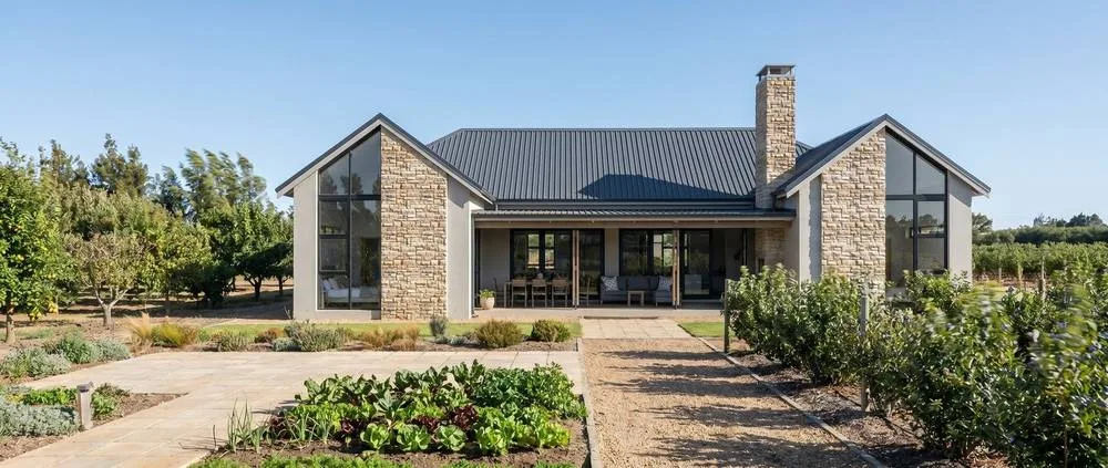 House Type 01, the Barn-Form, arrival side: a sheltered porch between two stone-clad gables, a paved approach and a vegetable garden.